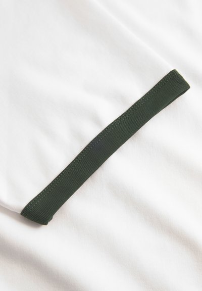 Tessuto bianco con una finitura elastica verde scuro. La texture è morbida e l'accento verde contrasta con il materiale bianco liscio.