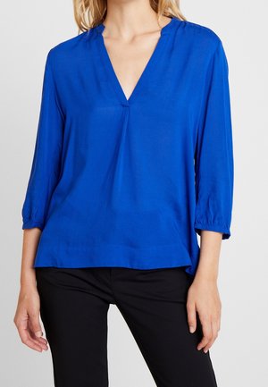 Femme portant un chemisier bleu roi à col en V avec des manches trois-quarts, associé à un pantalon noir ajusté sur un fond uni.