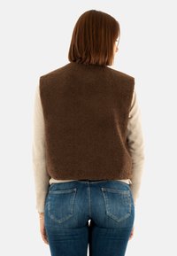 Gilet di pile marrone con collo alto, indossato sopra un maglione leggero, abbinato a jeans blu con tasche posteriori e una leggera sbiaditura.