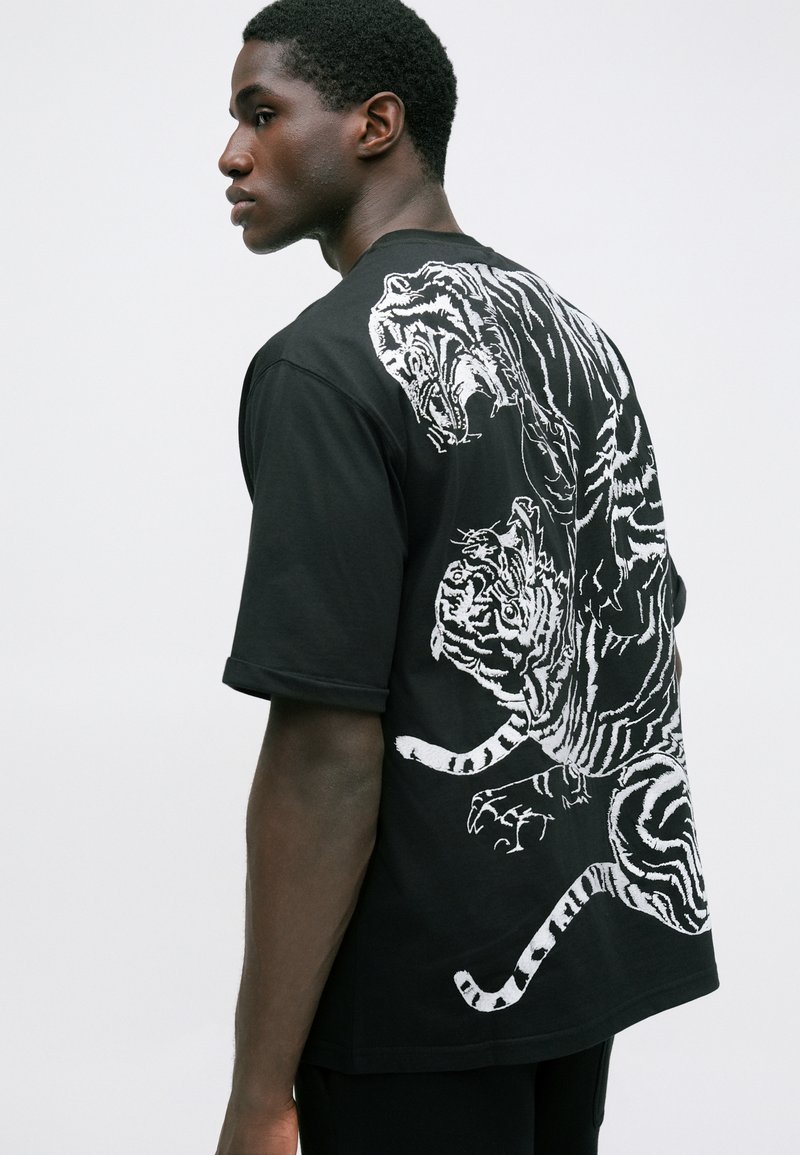 Jeune homme portant un t-shirt noir avec un grand motif de tigre blanc au dos, se tenant devant un fond clair, regardant sur le côté.