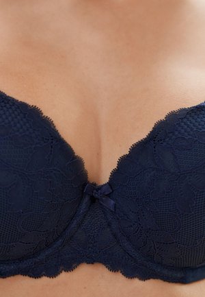 Tmavě modrá krajková podprsenka s květinovým vzorem a malou středovou mašlí, nošená na holé kůži, zobrazující přirozený výstřih.