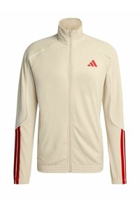 Giacca beige con cerniera integrale, collo alto, logo rosso sul petto sinistro e due strisce rosse che percorrono tutta la lunghezza di ogni manica.