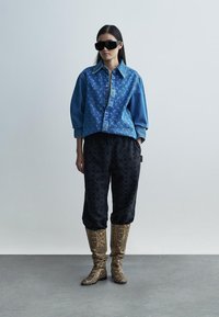Denim shirt with a swirl pattern, black patterned pants, and knee-high snake print boots. Model wears large black sunglasses.