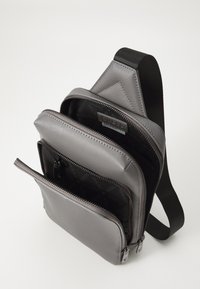 Grå syntetisk sling bag med flere glidelåskompartimenter. Har et strømlinjeformet design, slitesterkt materiale og justerbar stropp. Innsiden har et logomønster.
