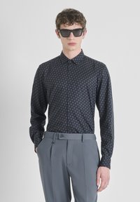 Camicetta nera a maniche lunghe con motivo a rombi, abbinata a pantaloni grigi su misura. Modello che indossa occhiali da sole scuri a forma quadrata.