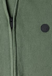 Gros plan d'un tissu tricoté côtelé vert avec une fermeture éclair noire partiellement fermée et un patch rond noir avec logo.