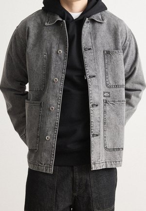 Mann trägt eine offene graue Jeansjacke mit großen Fronttaschen über einem schwarzen Kapuzenpullover und dunkler Jeans.