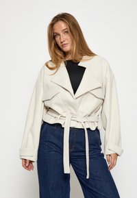 Chaqueta de crema con cinturón, cuello sobredimensionado, bolsillos con solapa y mangas anchas; combinada con pantalones de mezclilla azul oscuro. Tejido suave y estructurado.