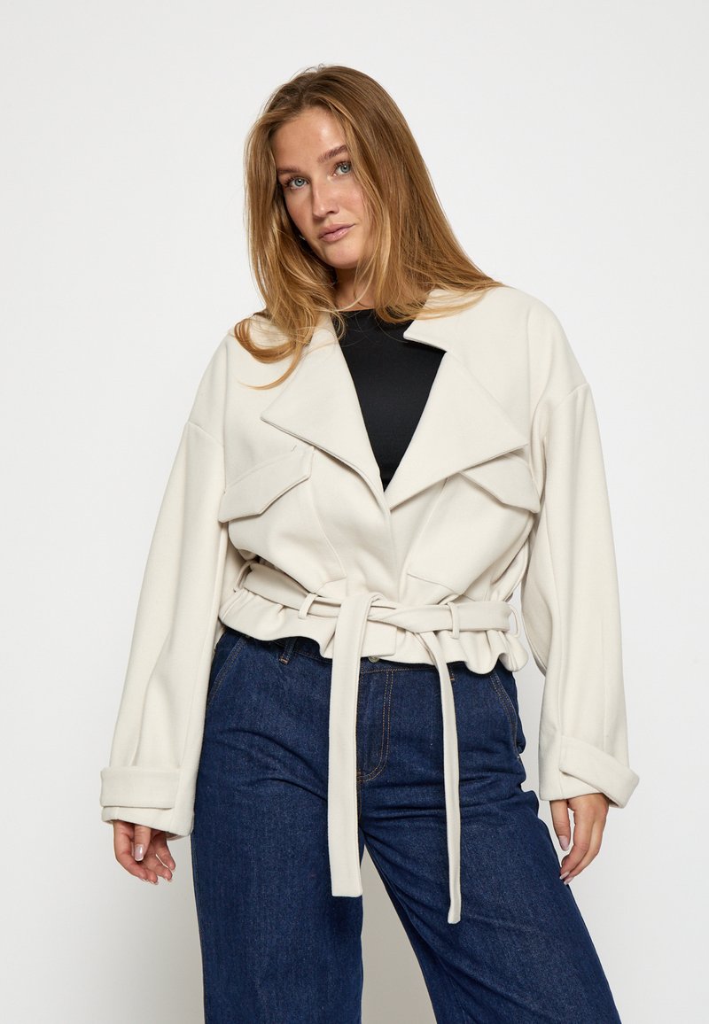 Chaqueta de crema con cinturón, cuello sobredimensionado, bolsillos con solapa y mangas anchas; combinada con pantalones de mezclilla azul oscuro. Tejido suave y estructurado.