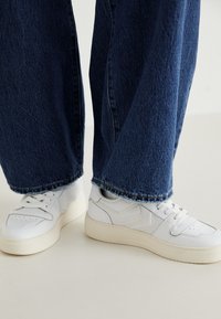 Osoba v širokých modrých džínách a bílých teniskách stojí na světlé podlaze před jednobarevným pozadím.