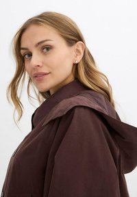 Donna con capelli castano chiaro che indossa orecchini a cerchio dorati e un cappotto con cappuccio marrone scuro, che guarda leggermente oltre la spalla contro uno sfondo semplice.