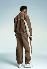 Brown lightweight jacket with white accents and a drawstring hem, paired with matching pants featuring side stripes. Worn with white sneakers.