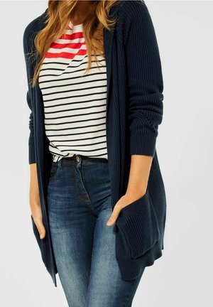 Kobieta ubrana w granatowy, prążkowany kardigan na guziki, na białej koszuli z czarnymi i czerwonymi paskami, w połączeniu z wyblakłymi, niebieskimi dżinsami.