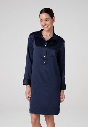 Donna sorridente che indossa un vestito camicia in raso blu navy fino al ginocchio con maniche lunghe, pattina con bottoni frontali e taschino sul petto.