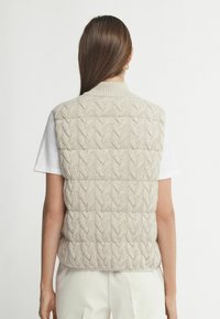 Beige gestrickte Weste mit Kabelstrickmuster, hohem Kragen und ärmellosem Design. Getragen über einem weißen Hemd mit strukturierter Oberfläche.