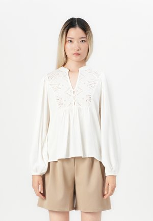 Blouse blanche à manches longues avec un col en V, broderie à œillets sur le haut et détail de bouton à l'avant. Tissu texturé avec une coupe fluide.