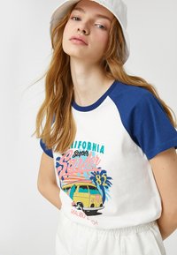 Vit och blå raglan t-shirt med en färgglad grafik av en vintagebuss och text, tillverkad av mjukt bomullsmaterial, lämplig för avslappnad användning.