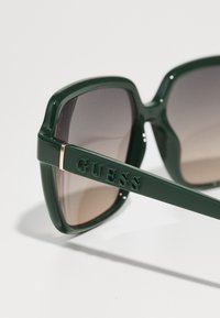 Zielone, oversize okulary przeciwsłoneczne z gradientowymi brązowymi soczewkami, o matowym wykończeniu i wytłoczonym logo "GUESS" na zauszniku.