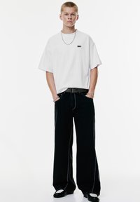 Hvid oversized t-shirt med lille sort logo, parret med sorte brede bukser med kontrastfarvet hvid syning og et tekstureret bælte.