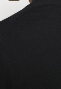 Svart stickad plagg med en texturerad yta, med rundad halsringning och synlig sömsdetaljering på axelområdet.