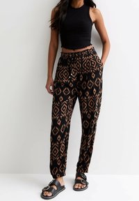 Femme portant un crop top sans manches noir, un pantalon ample à motifs noirs et bruns, et des sandales noires à boucle, se tenant devant un mur blanc uni.