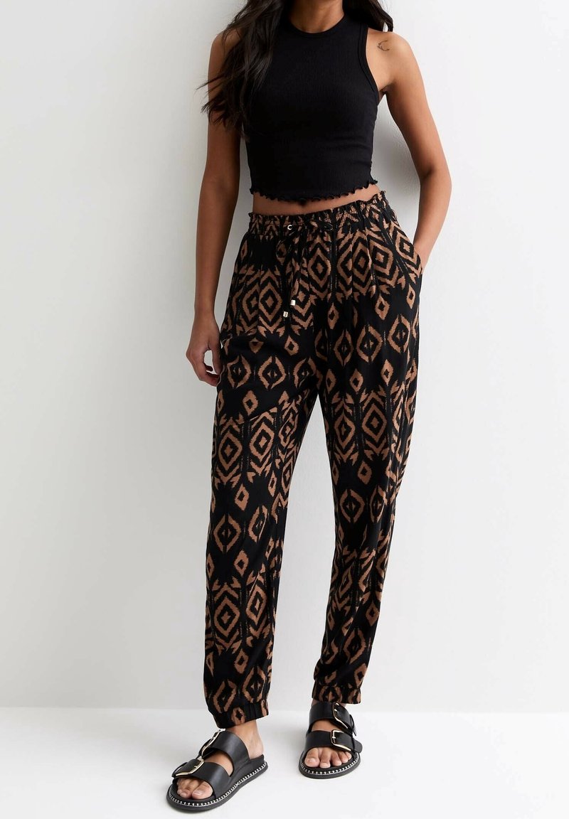 Femme portant un crop top sans manches noir, un pantalon ample à motifs noirs et bruns, et des sandales noires à boucle, se tenant devant un mur blanc uni.