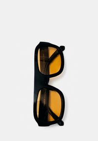 Lunettes de soleil à monture noire avec des verres rectangulaires couleur ambre reposant sur une surface blanche.