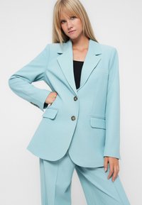 Svetlomodrý blazer s klopou, dvoma prednými vreckami a dvojitým zapínaním. Hladká textúra, prispôsobený strih, ktorý ladí s nohavicami v rovnakom odtieni.