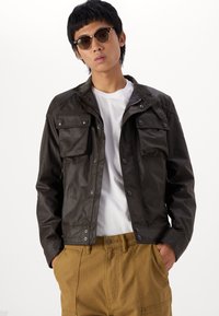 Σκούρο καφέ, ελαφρύ μπουφάν με κουμπιά snap και δύο τσέπες στο στήθος, συνδυασμένο με λευκό πουκάμισο και χακί παντελόνι.