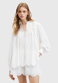 Blusa bianca con maniche a palloncino, design arricciato, chiusura a bottoni e orlo scallop. Realizzata in materiale leggero, adatta per abbigliamento casual.