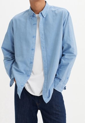 Mand iført lyseblå skjorte med knapper over hvid t-shirt og mørkeblå jeans, hænderne i lommerne, stående mod hvid baggrund.