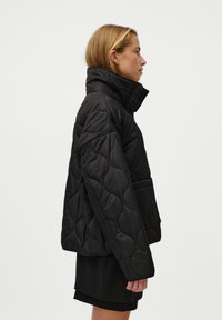 Black quilted jacket with a high collar, side pockets, and a boxy fit. Fabric has a smooth texture with a slight sheen.