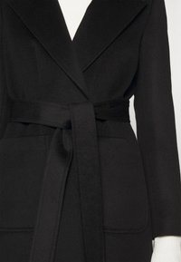 Wełniany płaszcz z czarnym paskiem i szerokimi klapami, noszony na białej prążkowanej bluzce z długim rękawem, ukazujący górną część tułowia i ramiona.