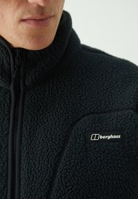 Giacca in pile nera con chiusura a zip, tessuto testurizzato e cuciture a contrasto. Presenta un patch con logo sul lato sinistro superiore.