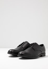 Scarpe da uomo in pelle nera con finitura liscia, stringhe sottili, punta rotonda e tacco basso; design minimalista senza marchi visibili.