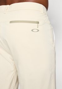 Beige athletic shorts with a zippered back pocket. Features a subtle logo on the side. Material appears lightweight and smooth.