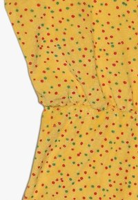 Tejido amarillo con un acabado texturizado, cubierto de pequeños lunares rojos y verdes. Presenta una cintura fruncida para dar forma.