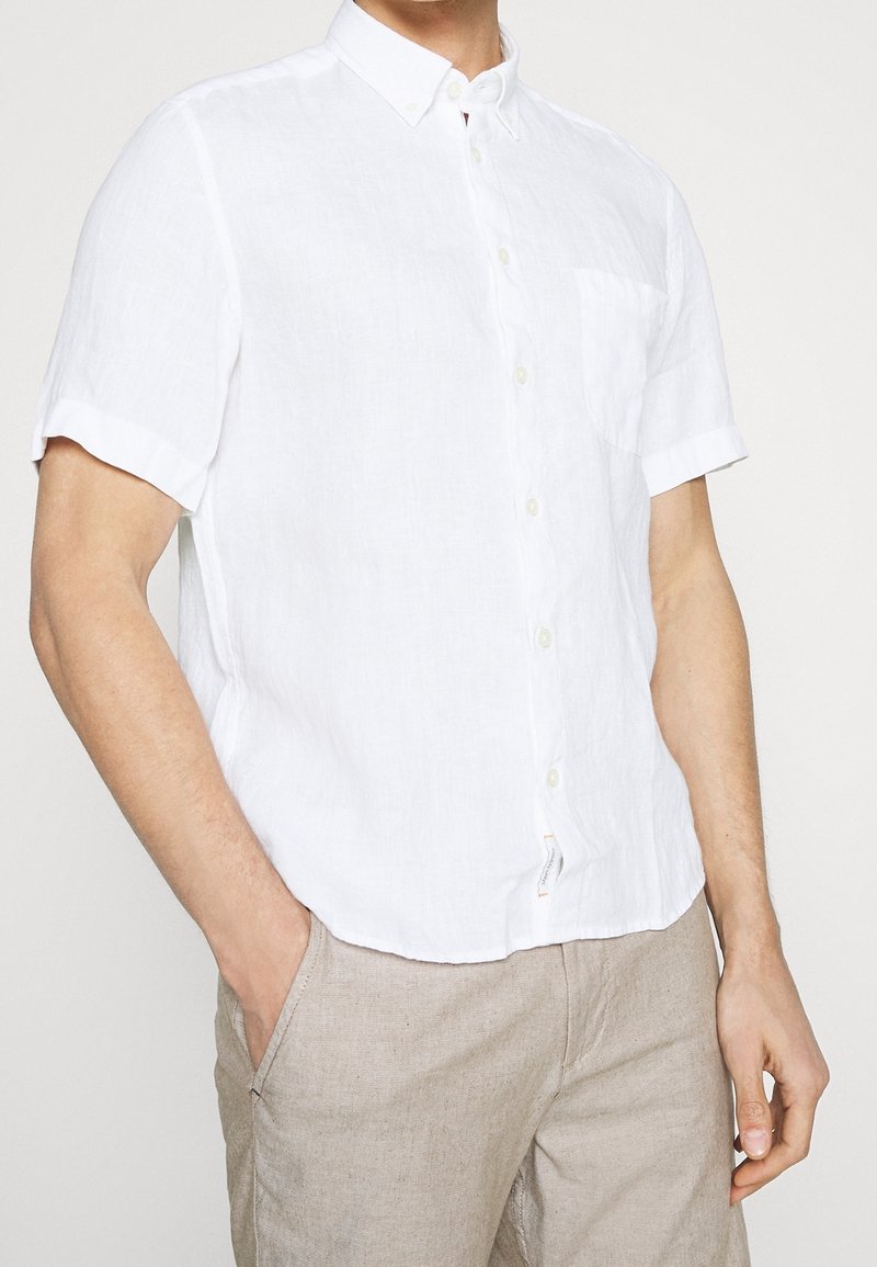 Camicia bianca di lino a maniche corte con colletto button-down, taschino sul petto e vestibilità rilassata. Indossata con pantaloni beige chiaro.