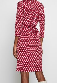 Punainen mekko, jossa on valkoinen timanttikuvio, kolmivartiset hihat ja takana sidottu vyötärö. Sileä kangas ja istuva muotoilu.