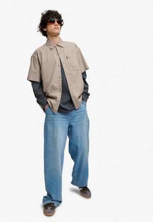 Ung person med solbriller, beige kortærmet skjorte over mørk langærmet skjorte, løse blå jeans og sneakers med hænderne i lommerne.