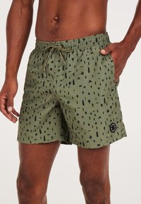 Zielone kąpielówki z czarnym abstrakcyjnym wzorem, elastyczny pas z sznurkiem do regulacji, boczne kieszenie oraz naszywka z logo na dolnym brzegu.