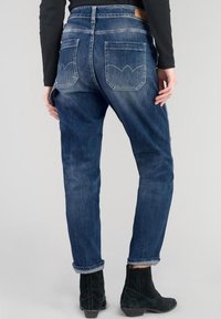 Jeans in denim blu scuro con vestibilità rilassata, dotati di due tasche posteriori con cuciture curve e orlo risvoltato. Indossati con stivaletti neri alla caviglia.