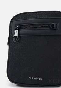 Zwarte schoudertas met een getextureerd logo-patroon, een ritszak aan de voorkant en een gemarkeerd label. Gemaakt van duurzame stof met een gladde afwerking.