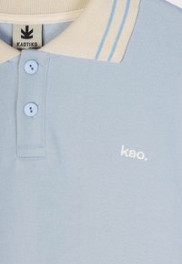 Svetlomodrá polo košeľa s béžovým golierom, obsahujúca dva gombíky a vyšitým logom "kao." v bielej farbe. Mäk-ká textúra, čistý dizajn so pruhmi.