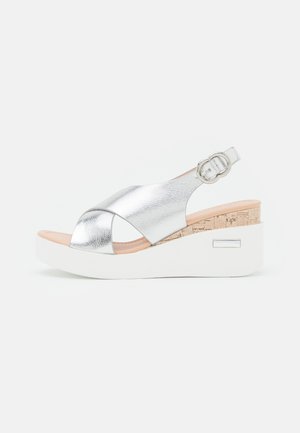 Zilverkleurige metallic sleehak sandaal met kruiselings gespen, verstelbare gesp, kurkdetail boven witte plateauzool op een effen achtergrond.
