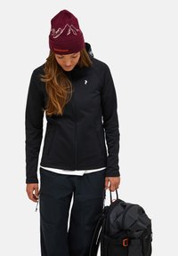 Men's black softshell jacket with zippered pockets, paired with dark pants and a maroon winter beanie featuring mountain graphics.