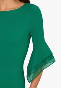 Abito verde con scollo rotondo e maniche a tre quarti, caratterizzato da volant trasparenti a strati sui polsi. Tessuto con una texture morbida.