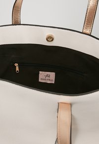 Bolso de tote blanco texturizado con acentos de hardware dorado, interior negro y un bolsillo con cremallera que presenta una etiqueta de la marca.