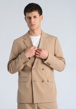 Ung mand, der justerer beige dobbeltradet jakkesæt, iført hvid skjorte, stående foran en lys grå baggrund.