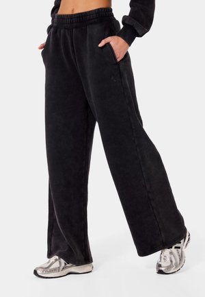 Osoba nosząca luźne czarne spodnie dresowe z elastycznym paskiem i kieszeniami, połączone ze srebrno-białymi butami sportowymi.